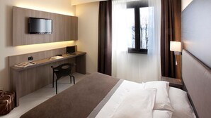 A hotel room with a bed, a desk, a chair, a TV mounted on the wall, and a window with curtains.