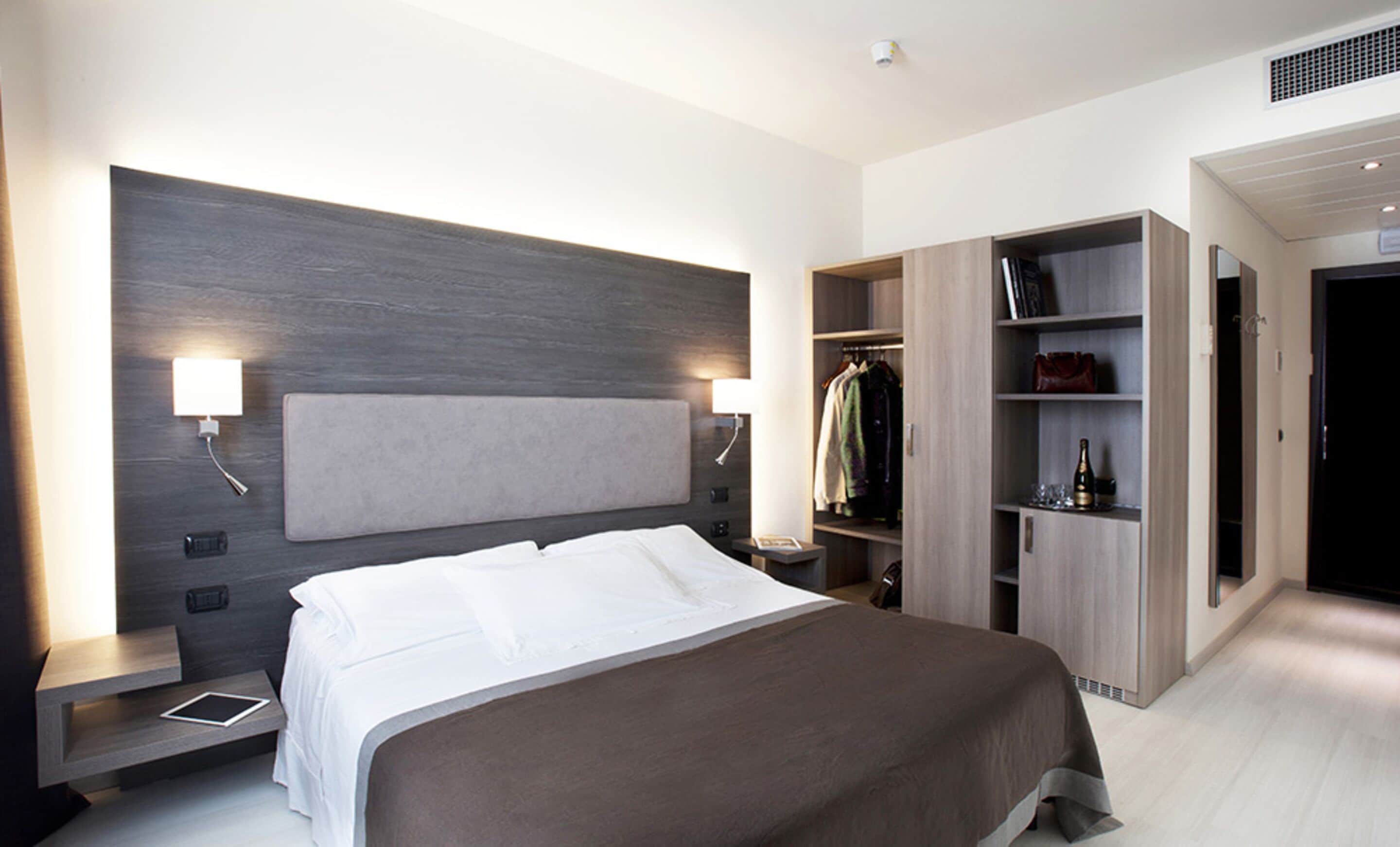 A modern hotel room with a large bed, bedside tables, a wardrobe, and a built-in shelf with a bottle and glasses.