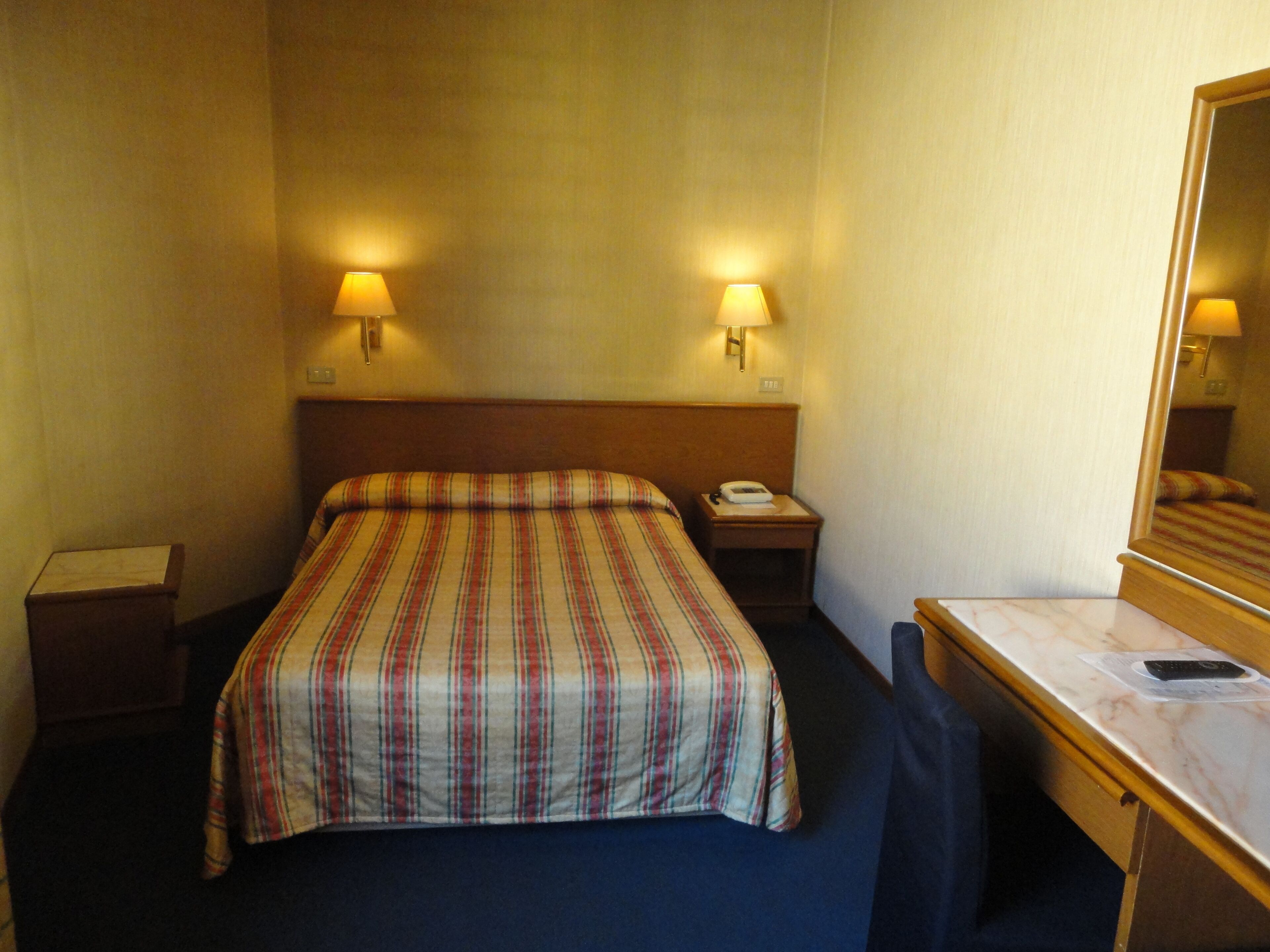 Een hotelkamer met een bed, nachtlampjes, een nachtkastje, een bureau en een spiegel.