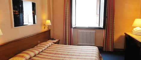 A hotel room with a bed, a window with curtains, a radiator, a mirror, and a desk with a lamp.