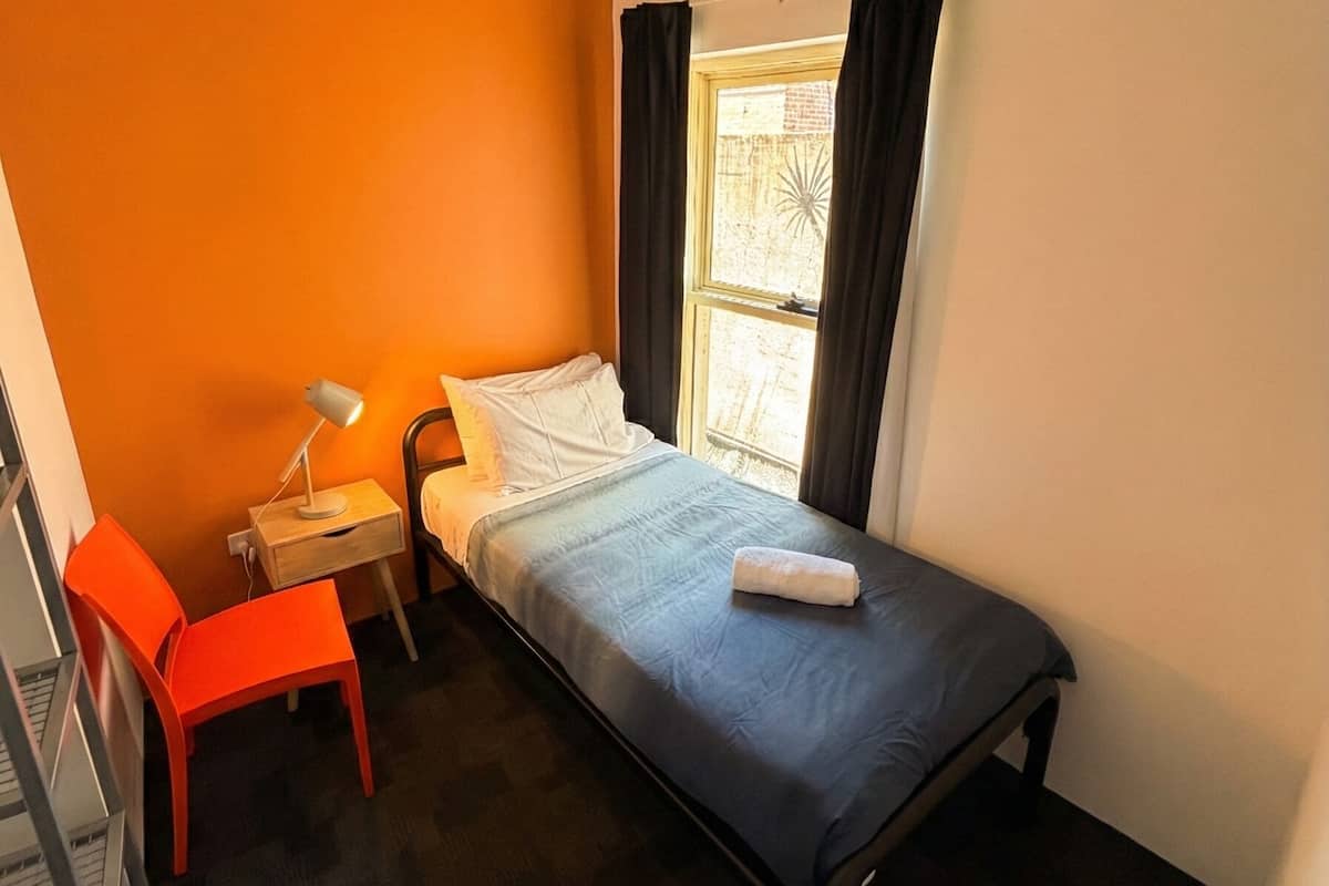 Una habitación pequeña con cama individual, pared naranja, un escritorio con silla, una ventana con cortinas y un radiador.