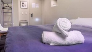 A neatly made bed with a rolled white towel on it, a purple bedspread, and a white wall with a small window and a framed picture.