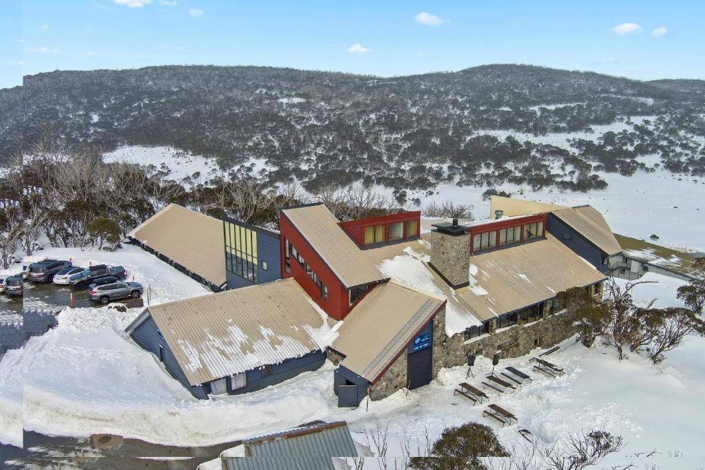 A building complex with multiple structures, including a red building with a sloped roof, a blue building with a flat roof, and a stone building with a flat roof. There is a parking area with several cars and a snow-covered landscape in the background.