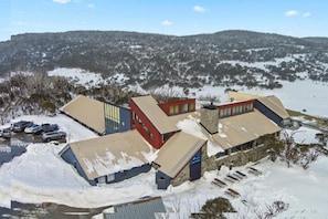 A building complex with multiple structures, including a red building with a sloped roof, a blue building with a flat roof, and a stone building with a flat roof. There is a parking area with several cars and a snow-covered landscape in the background. - Snowy Gums (Smiggin Holes)