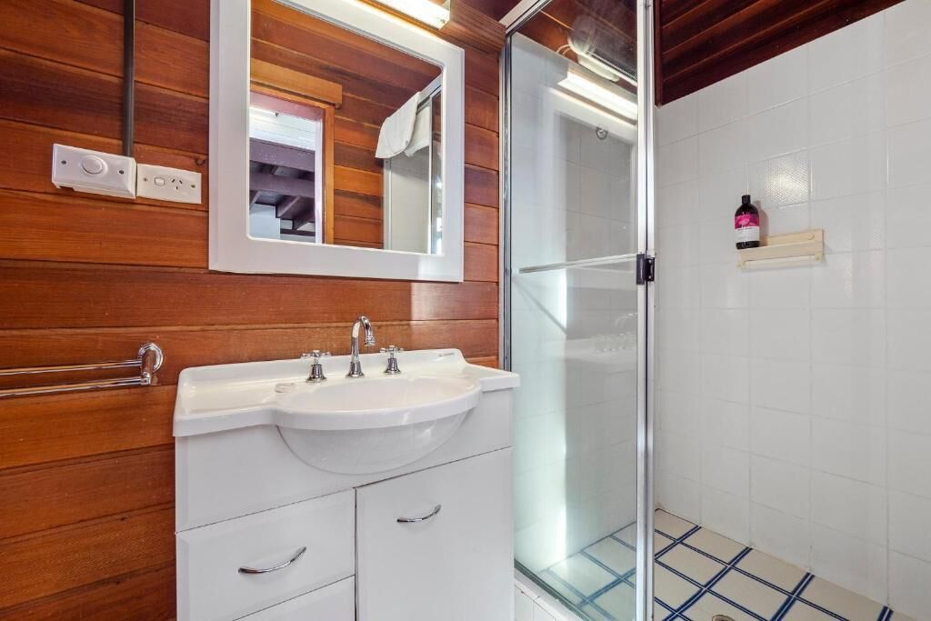 A modern bathroom with a wooden wall, a white sink with two faucets, a mirror, and a glass shower enclosure.