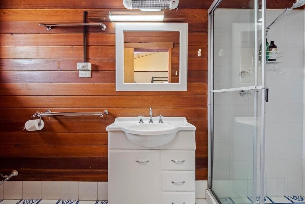 A bathroom with wooden panel walls, a white sink with two faucets, a mirror, and a glass shower enclosure.