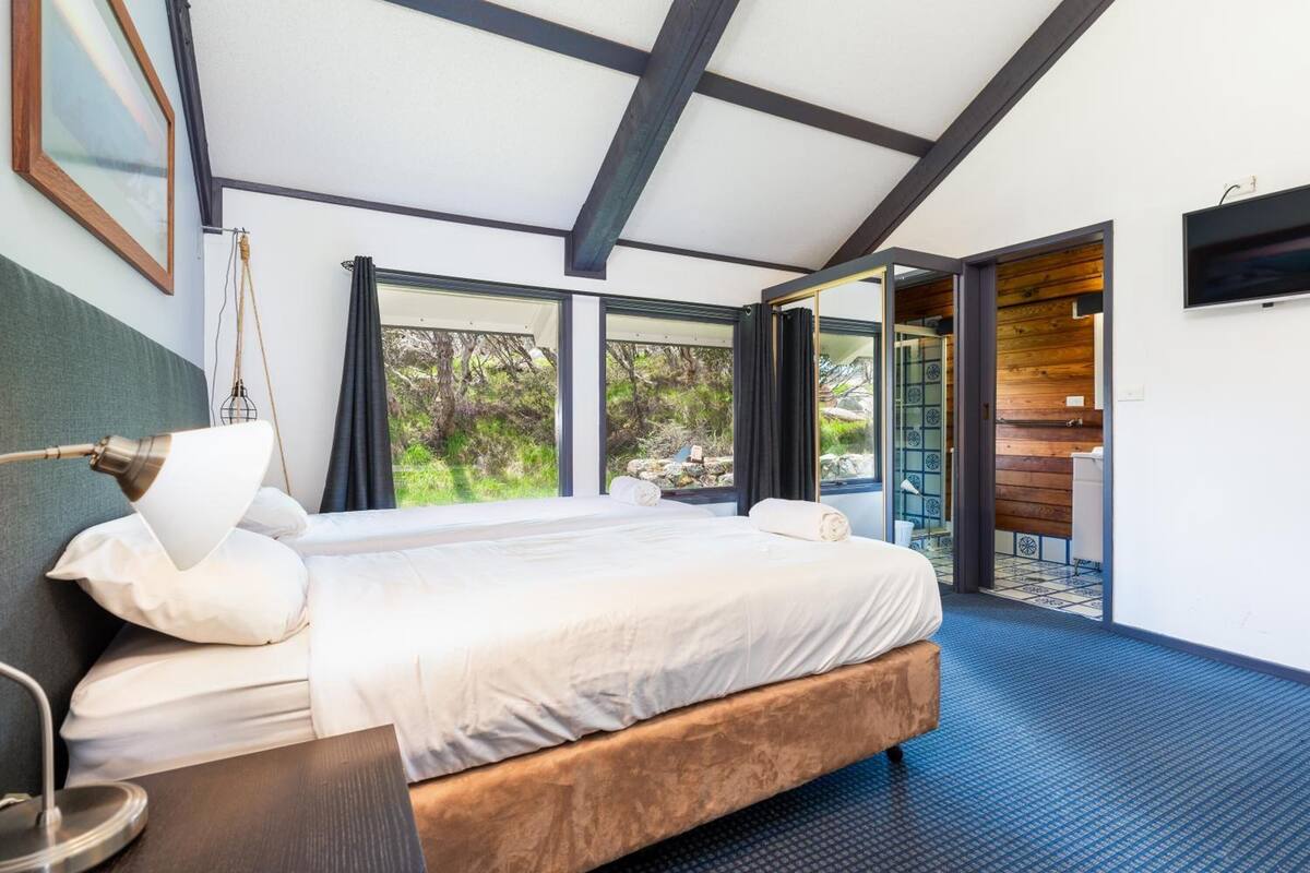 A modern bedroom with a large bed, a desk, and a view of the outdoors through large windows.