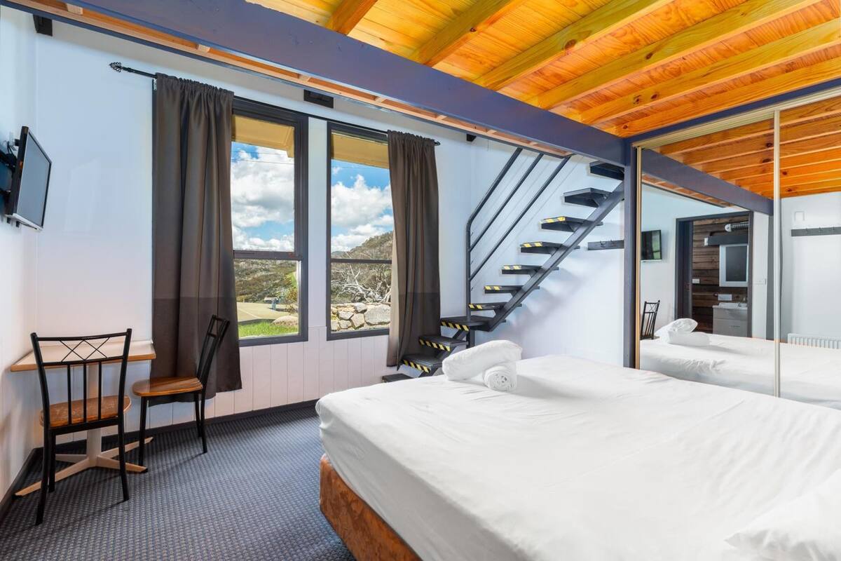 A hotel room with a large bed, a desk with a chair, a window with curtains, and a staircase leading to another room.