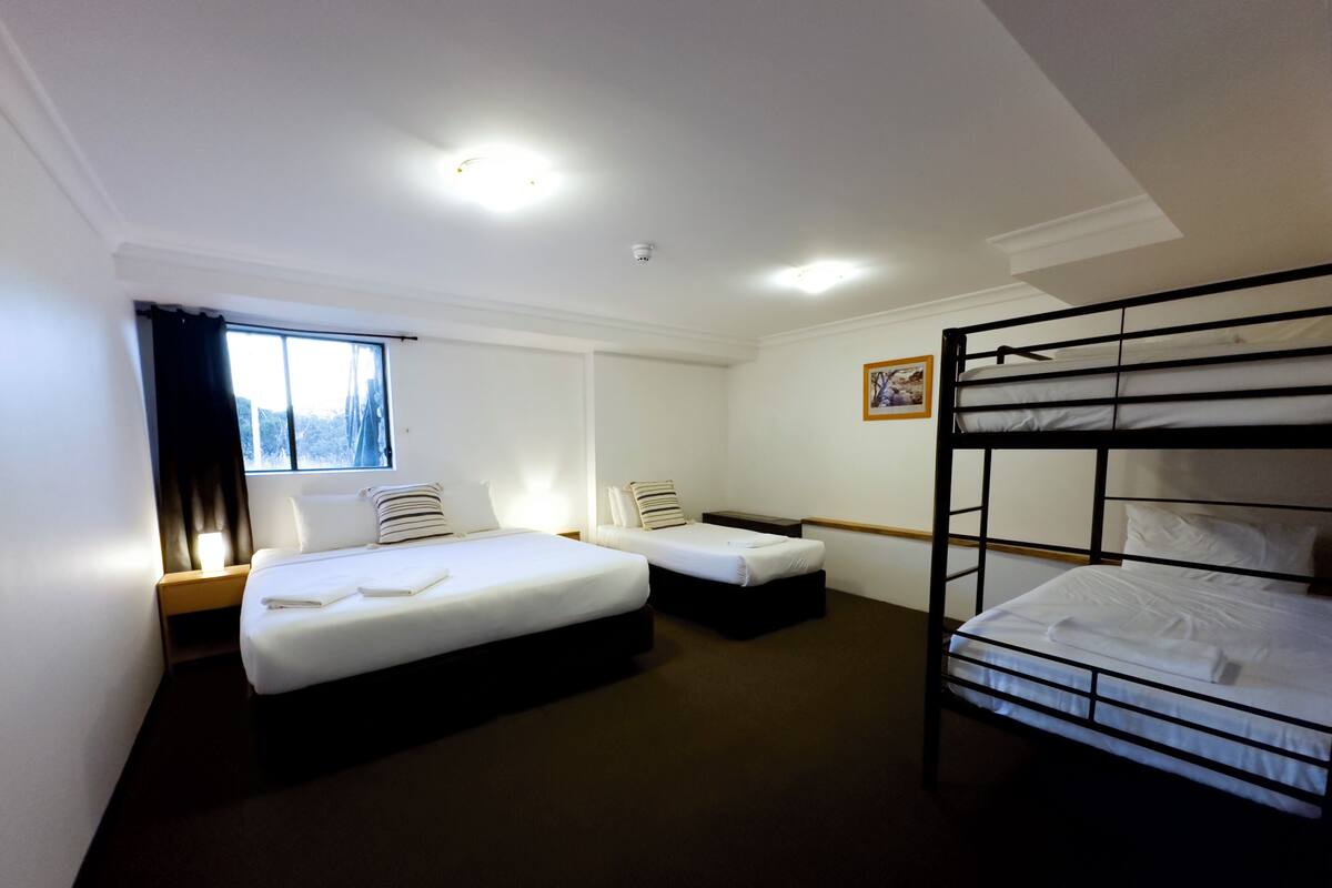 A hotel room with two single beds, a bunk bed, a window with curtains, a bedside table, and a wall-mounted picture.