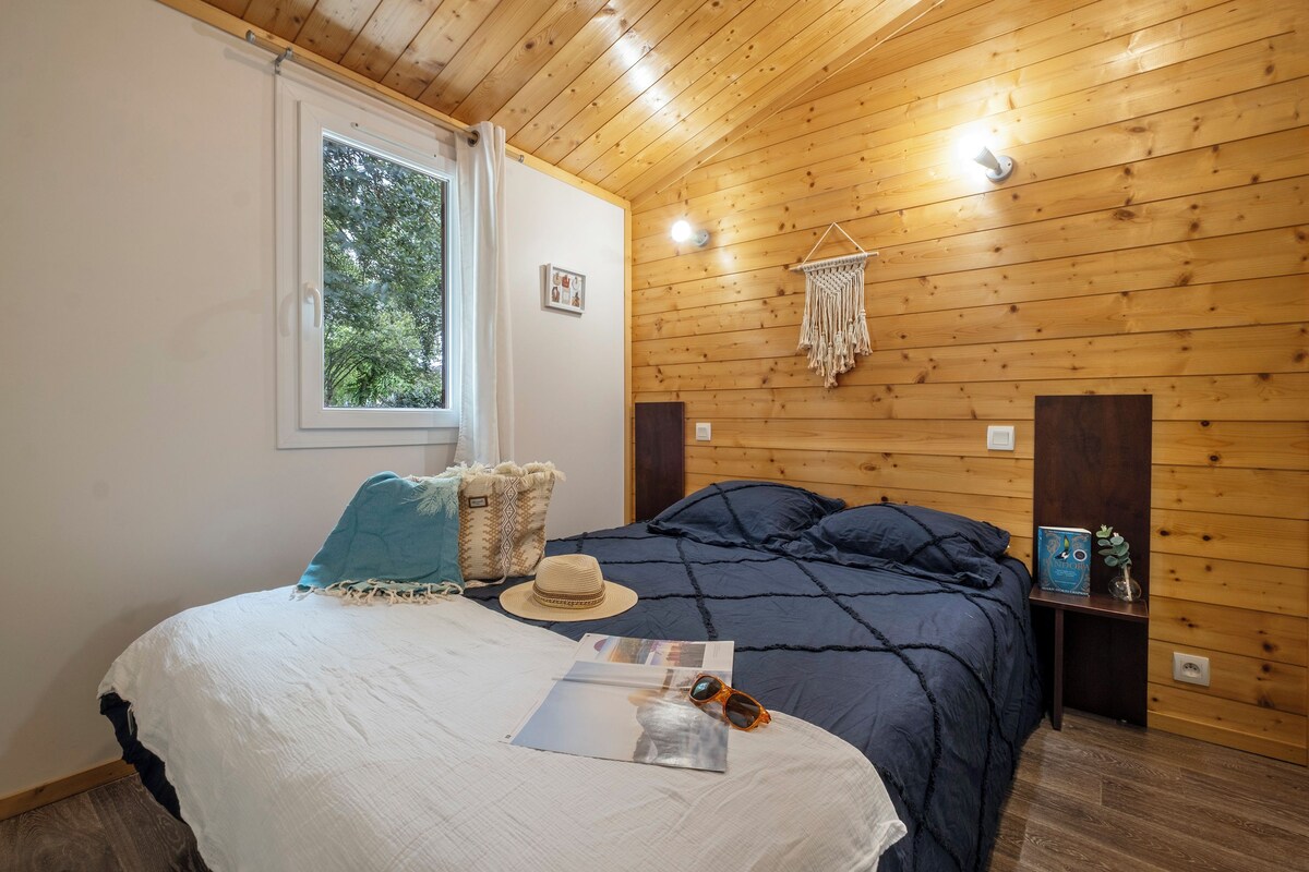 A wooden-paneled room with a bed, a window, a wall-mounted macrame decoration, and a small shelf with a plant.