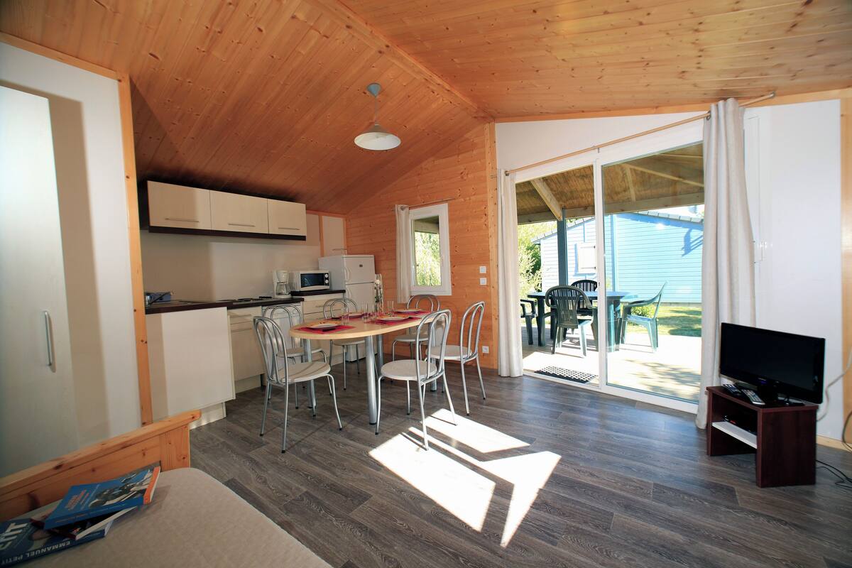 A compact, well-lit room with a wooden ceiling, a dining area with chairs and a table, a kitchenette, and a television on a stand.