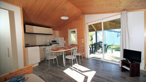 Una habitación compacta y bien iluminada con techo de madera, comedor con sillas y mesa, kitchenette y televisor sobre un mueble.