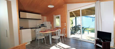 A compact, well-lit room with a wooden ceiling, a dining area with chairs and a table, a kitchenette, and a television on a stand.