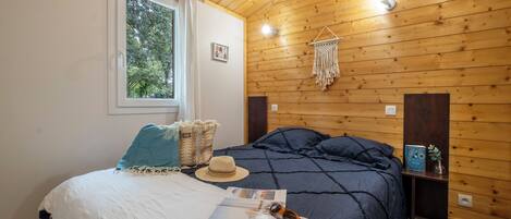 A wooden-paneled room with a bed, a window, a wall-mounted macrame decoration, and a small shelf with a plant.