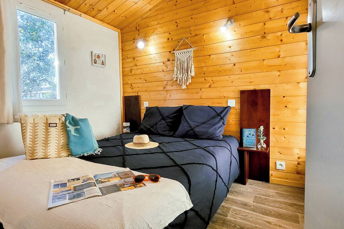 A cozy bedroom with a wooden ceiling, a large bed with a dark blue bedspread, a window with curtains, and a wall-mounted light fixture.