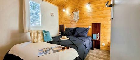 A cozy bedroom with a wooden ceiling, a large bed with a dark blue bedspread, a window with curtains, and a wall-mounted light fixture.