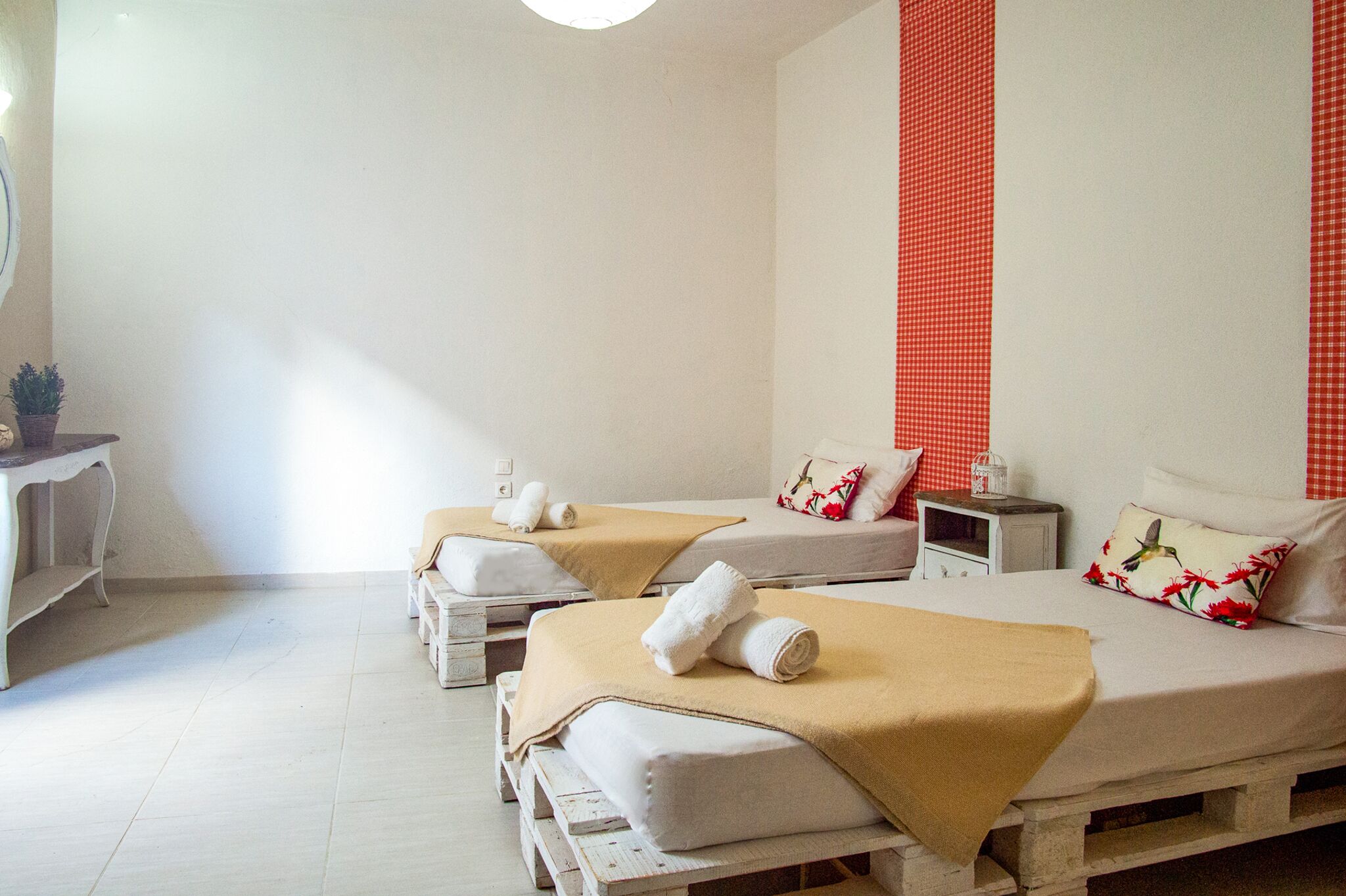 Habitación con dos camas, cada una con una manta color beige y almohadas blancas, una mesita de noche pequeña con una lámpara y una pared con diseño de cuadros rojos.