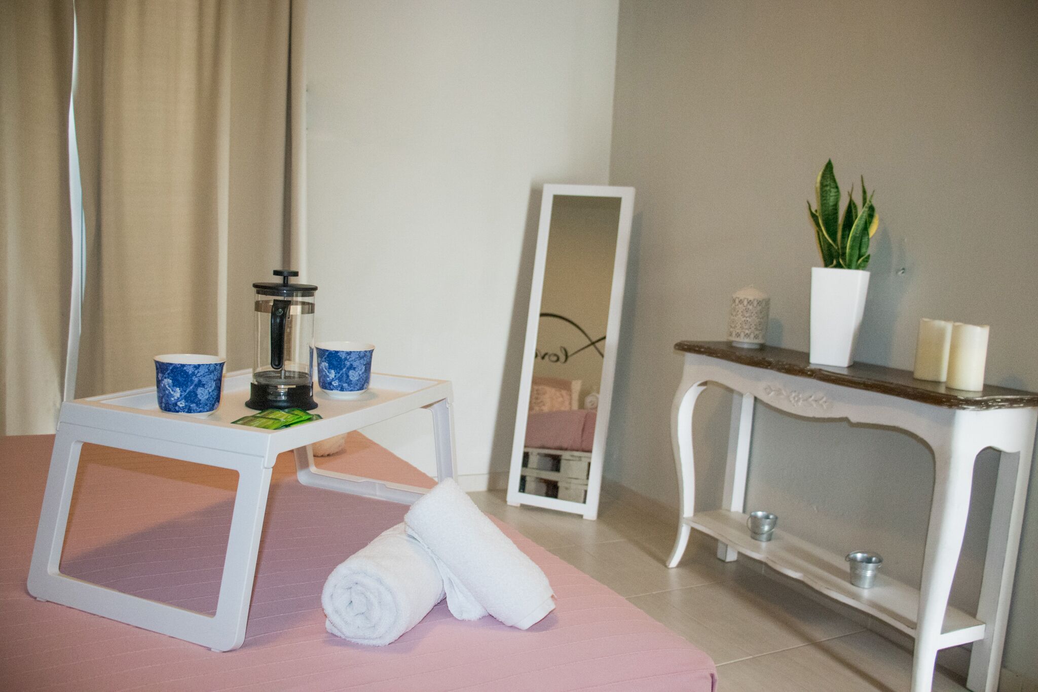 Una habitación con una camilla de masaje, una pequeña mesa blanca con cafetera, un espejo, una consola con una planta y velas.