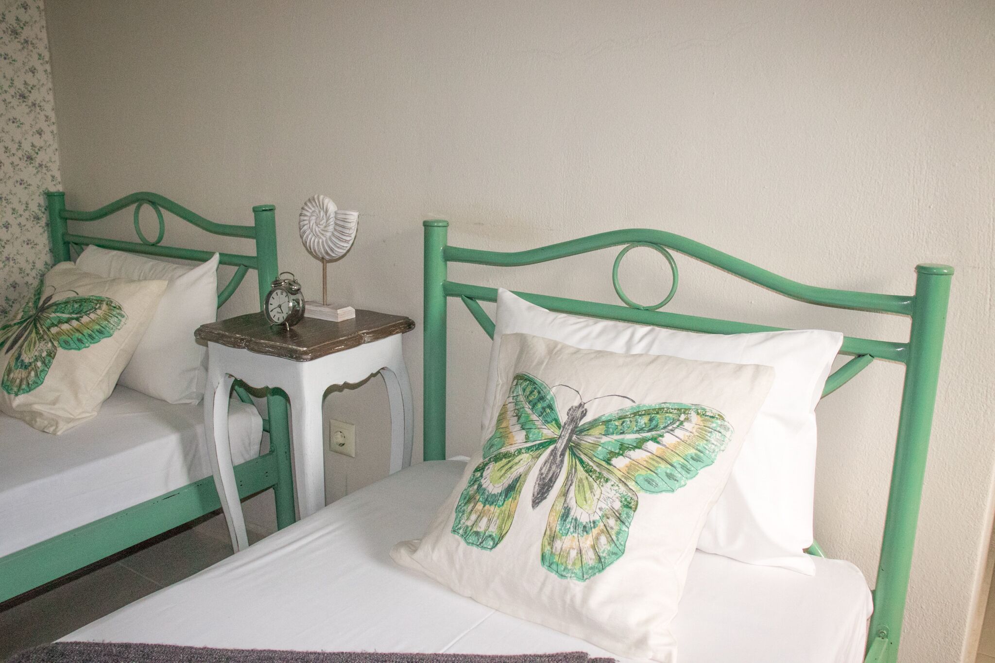 Una habitación con dos camas, cada una con cabecero verde y ropa de cama blanca. Hay una mesita de noche pequeña con un reloj y un adorno.