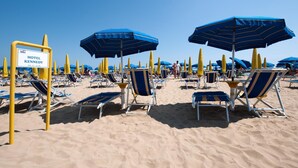Une plage bordée de rangées de transats bleus et blancs, de poteaux jaunes et de parasols bleus. On peut voir un panneau indiquant l’hôtel Kennedy.