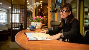 Un comptoir d’accueil avec une femme assise derrière, un comptoir en bois et un vase rempli de fleurs.