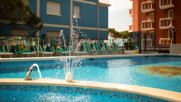 Une piscine avec une fontaine, des chaises longues vertes et un bâtiment en arrière-plan.