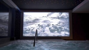 A spa room with a large screen displaying a snowy landscape, a hot tub, and a stone wall. - La Posada de Paco (Nijar)
