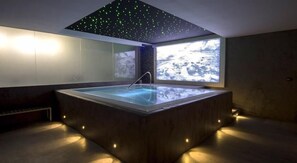 Un moderno spa con una bañera de hidromasaje rectangular, techo estrellado y un amplio ventanal con vistas a un paisaje rocoso.
