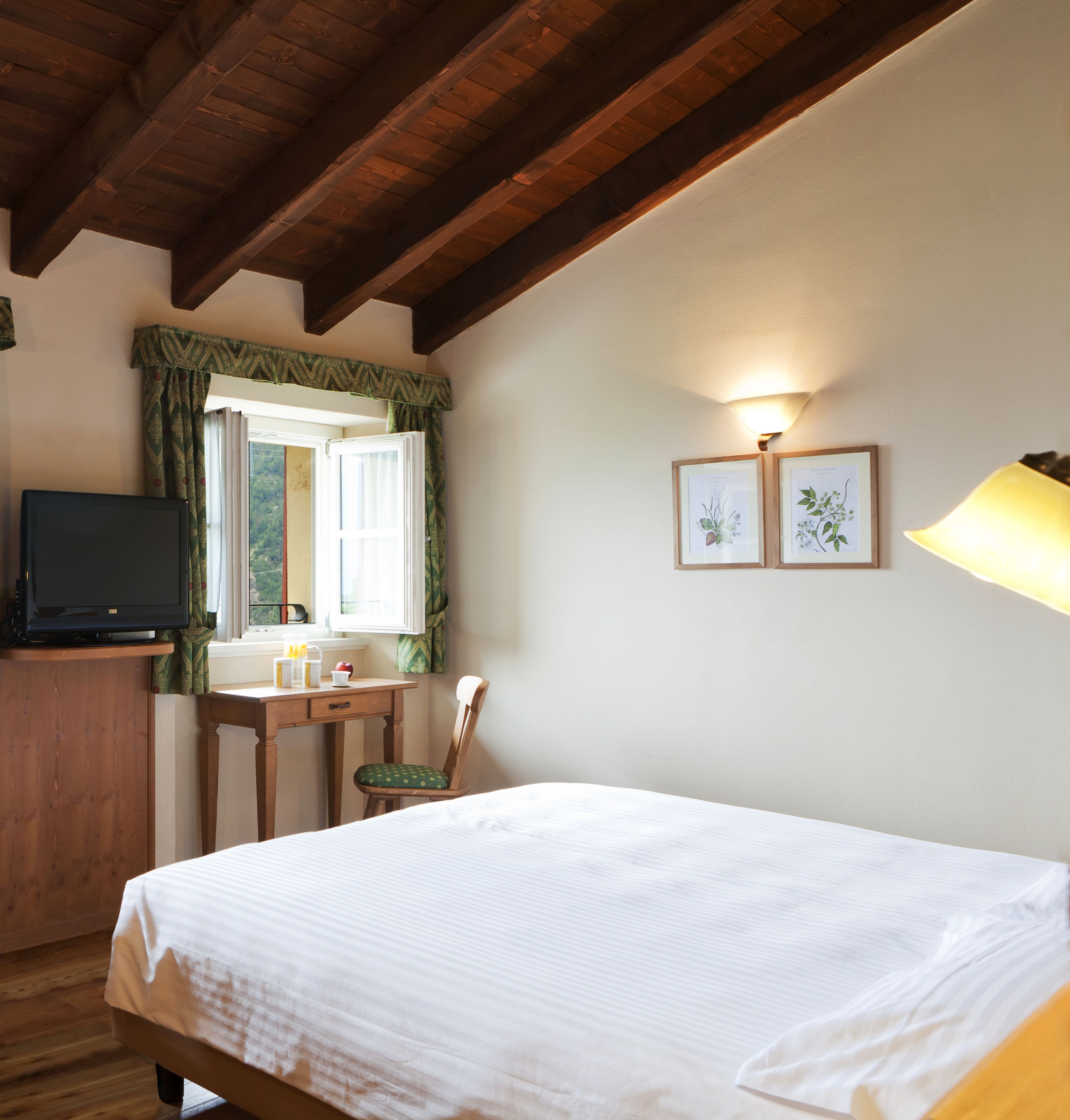 A bedroom with a wooden ceiling, a bed, a desk with a chair, a television, and two framed botanical prints on the wall.