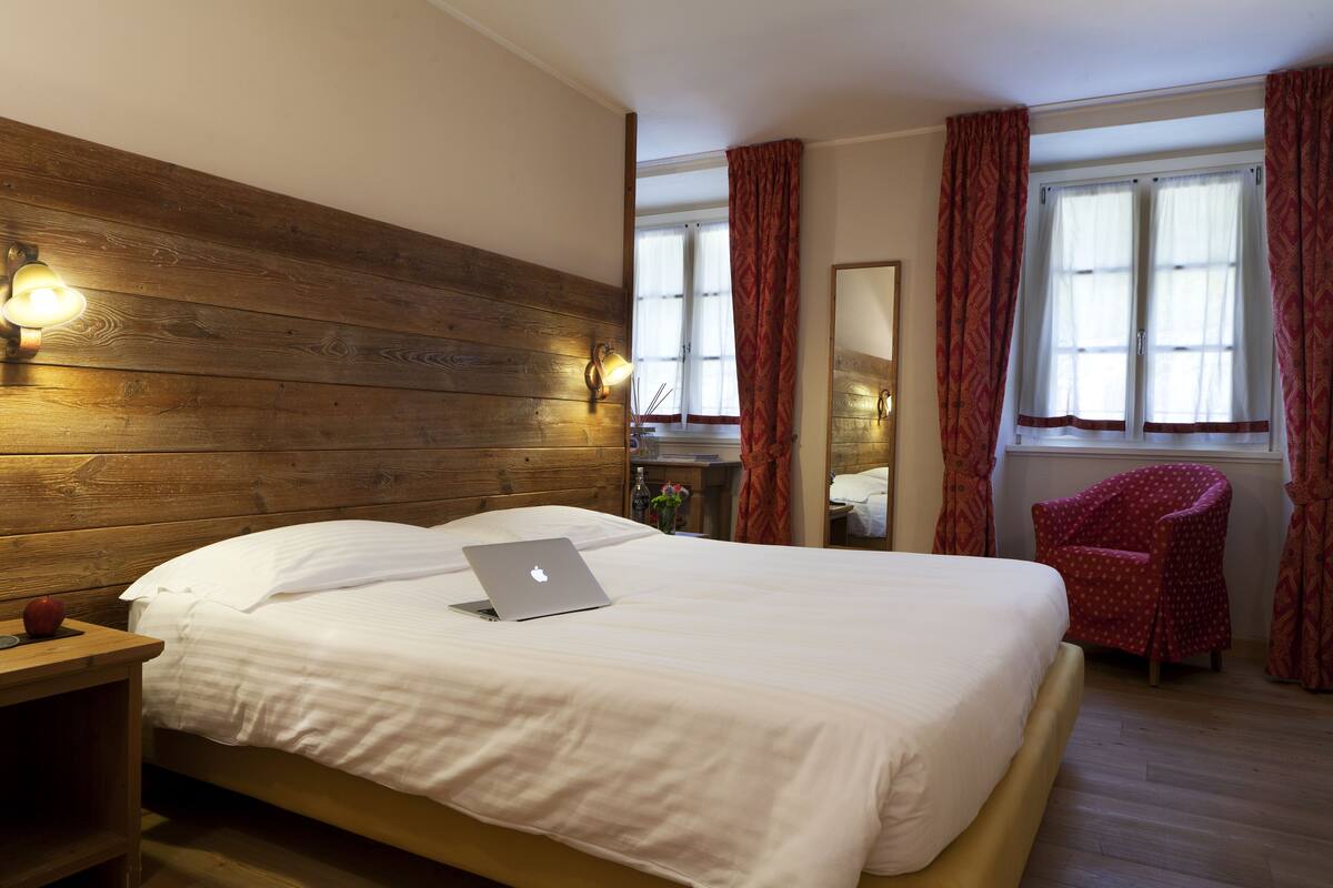 Ein ordentlich bezogenes Bett mit Laptop, Holz-Kopfteil und Stuhl am Fenster.