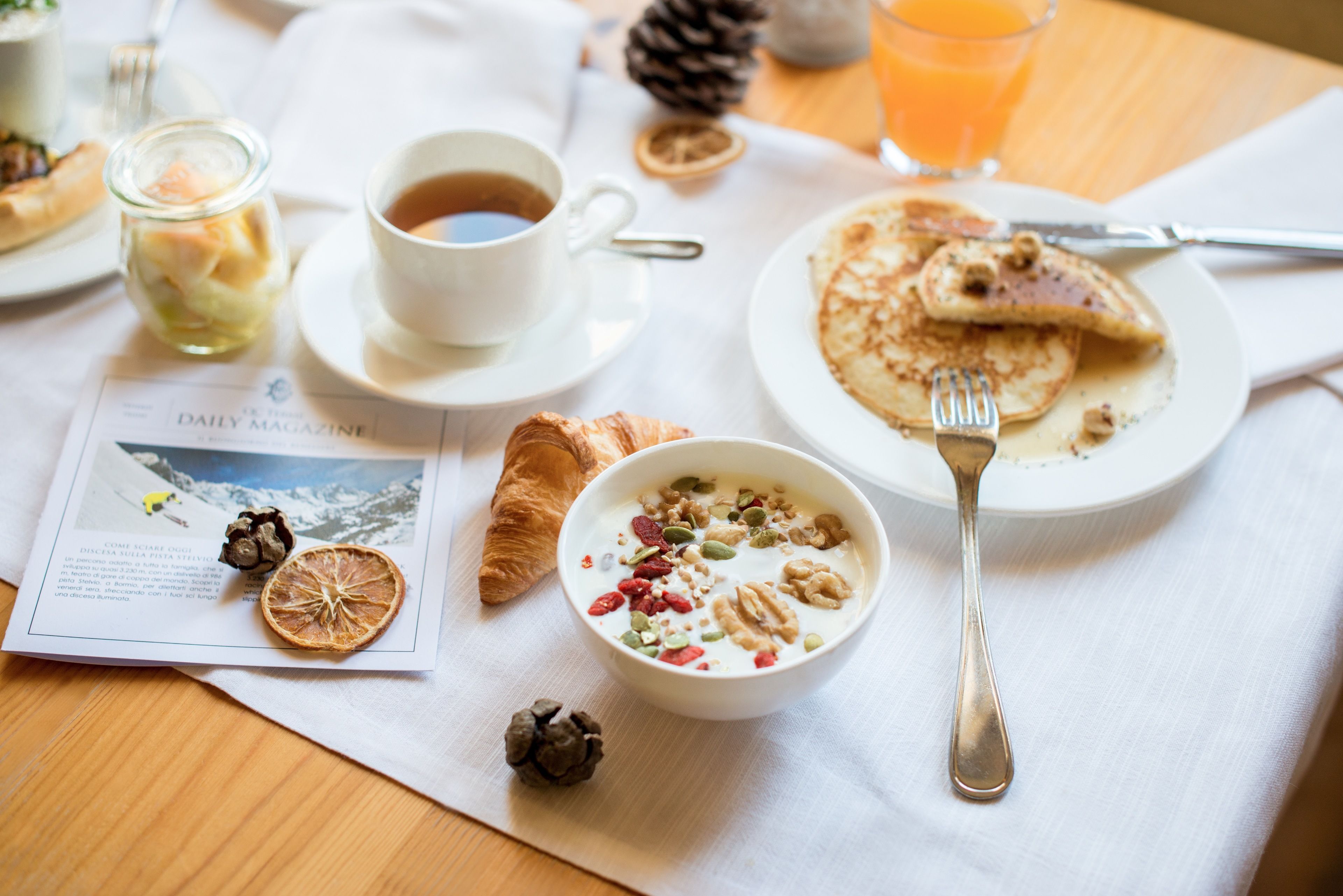 A breakfast setting with a croissant, a bowl of cereal, a cup of coffee, and a glass of orange juice on a table.