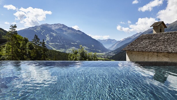 Une piscine aux eaux cristallines offrant une vue sur les montagnes, un petit bâtiment surmonté d’une croix et une végétation luxuriante.