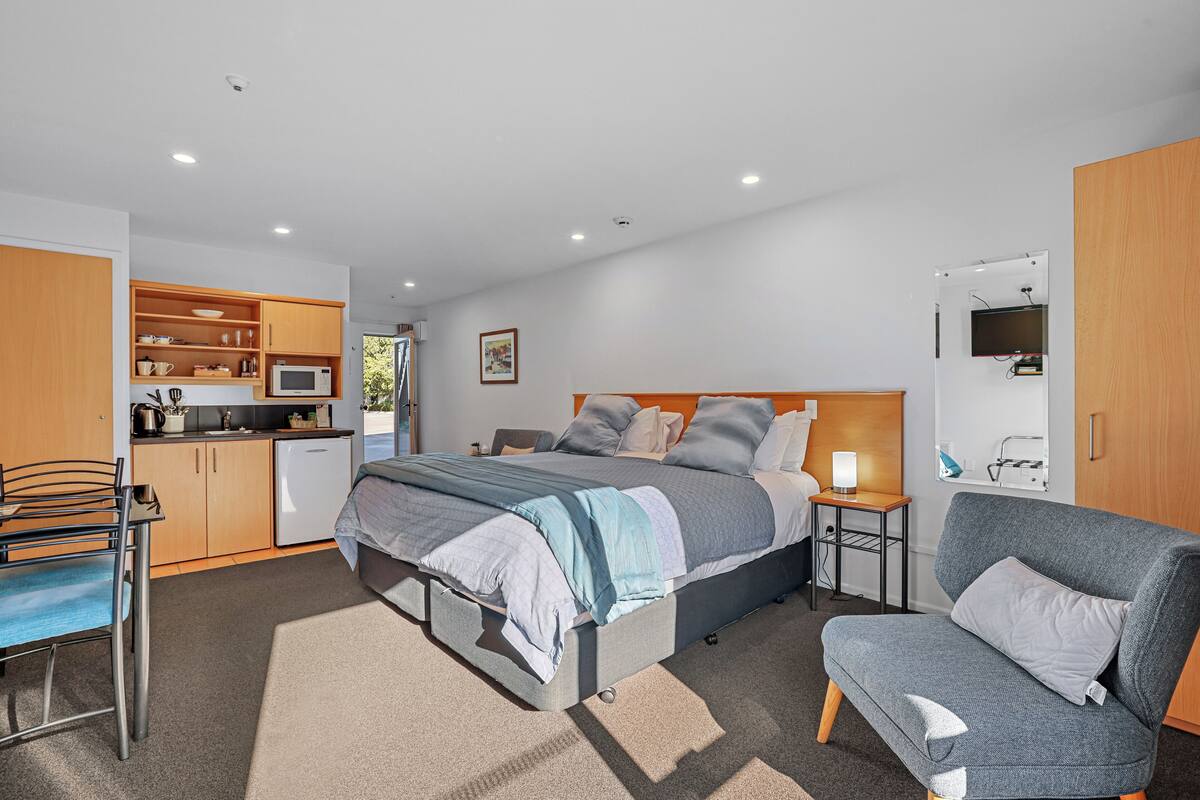A compact bedroom with a bed, bedside tables, a chair, a desk, and a kitchenette.