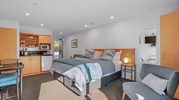 A compact bedroom with a bed, bedside tables, a chair, a desk, and a kitchenette.