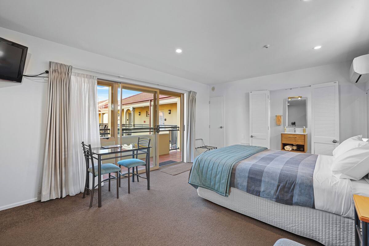 A bedroom with a bed, a desk with chairs, a TV, and a balcony with a view of buildings.