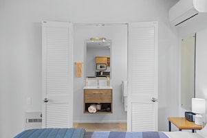 A hotel room with a bed, a bedside table with a lamp, a wooden cabinet with toiletries, and a mirror on the wall.