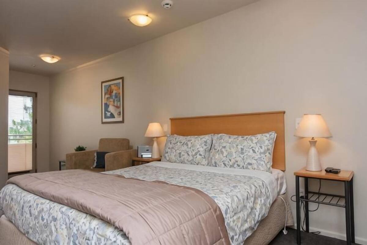 A bedroom with a large bed, two bedside tables with lamps, a chair, and a painting on the wall.