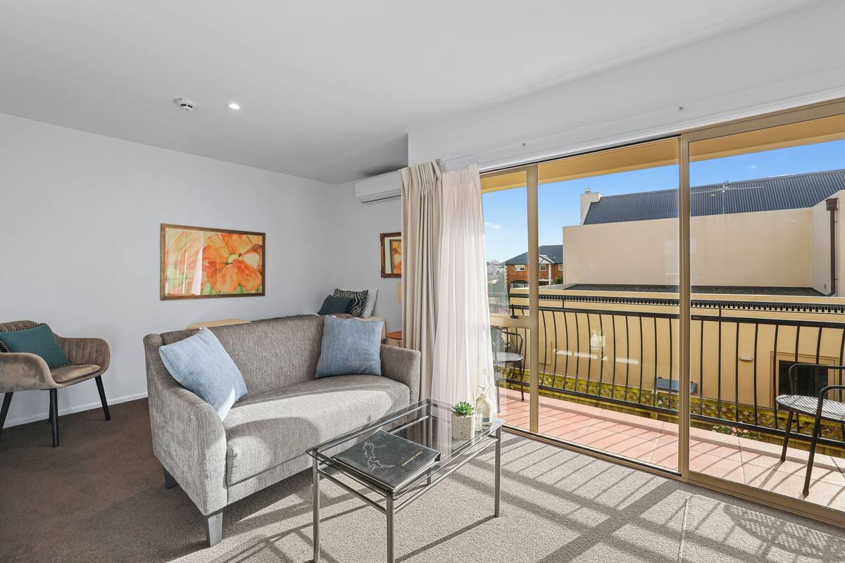 A modern living room with a grey sofa, a glass coffee table, and a view of a balcony with a railing.