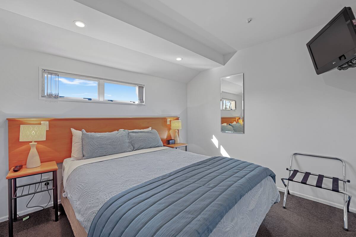 A modern bedroom with a large bed, two bedside tables, a nightstand, a window with blinds, a mounted TV, and a mirror on the wall.