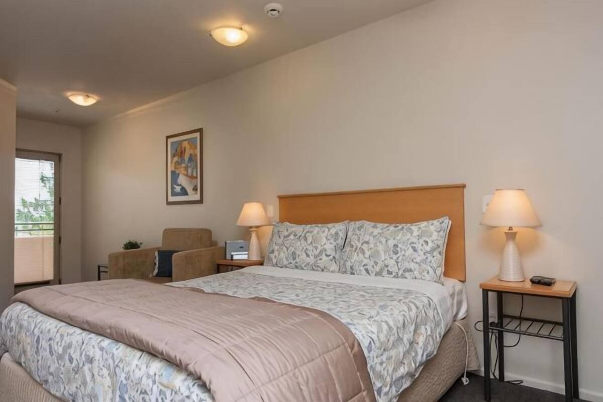 A hotel room with a bed, two bedside lamps, a chair, a small table, and a painting on the wall.