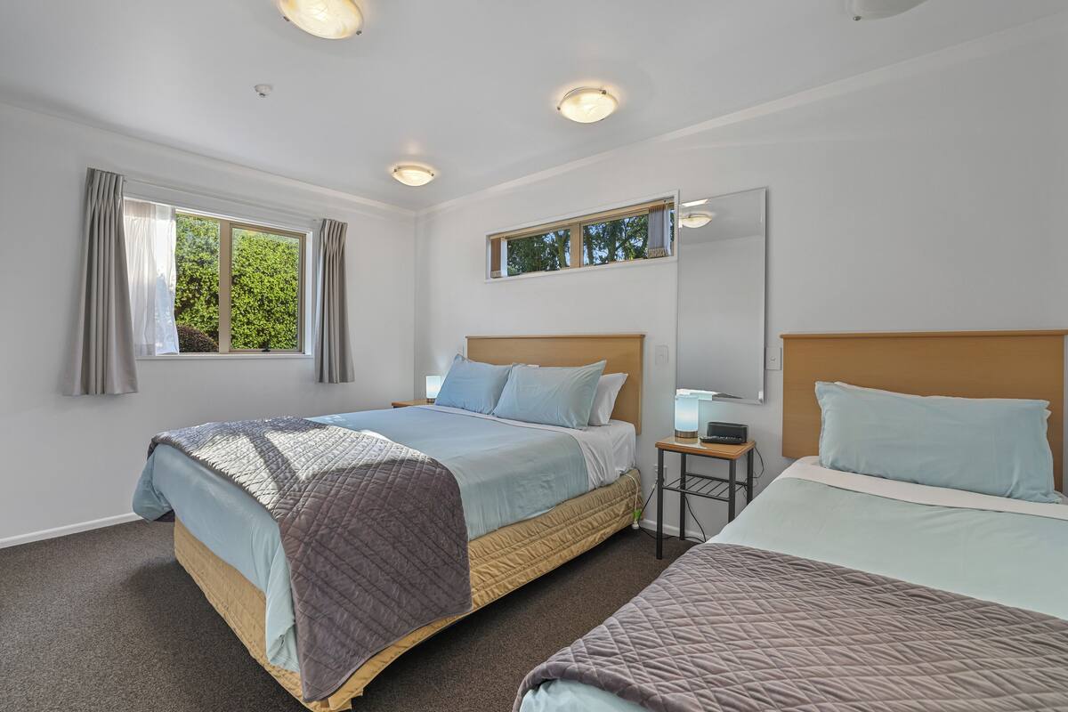 A hotel room with two beds, a small table with a lamp, a window with curtains, and a mirror on the wall.