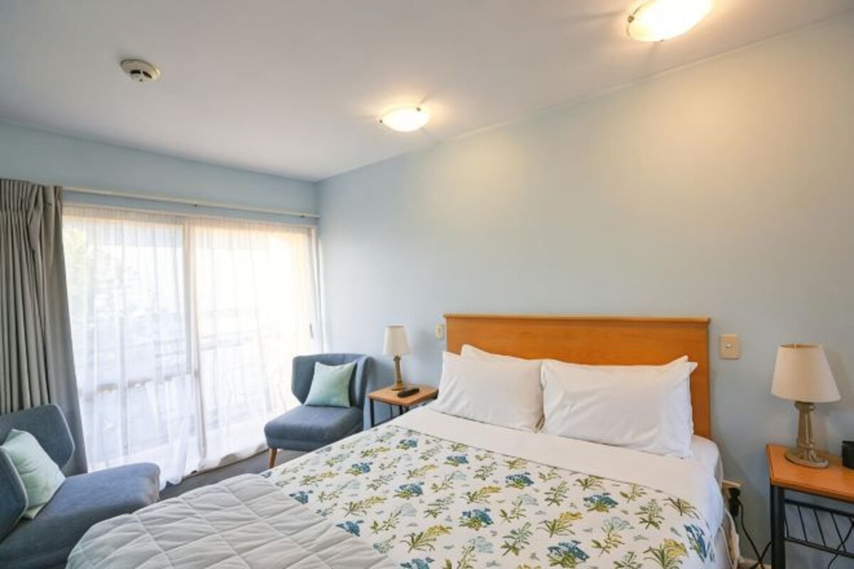 A hotel room with a bed, a chair, a nightstand, and a window with curtains.