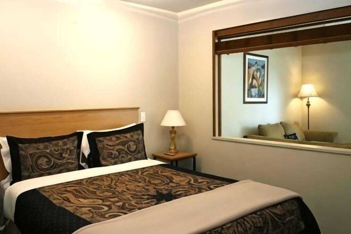 A hotel room with a bed, a nightstand with a lamp, a window with a view, and a painting on the wall.