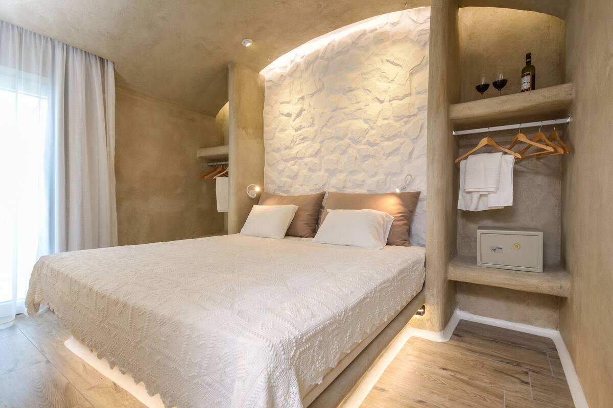 A bedroom with a large bed, a stone-textured wall, a wooden floor, a shelf with wine bottles, and a towel rack.