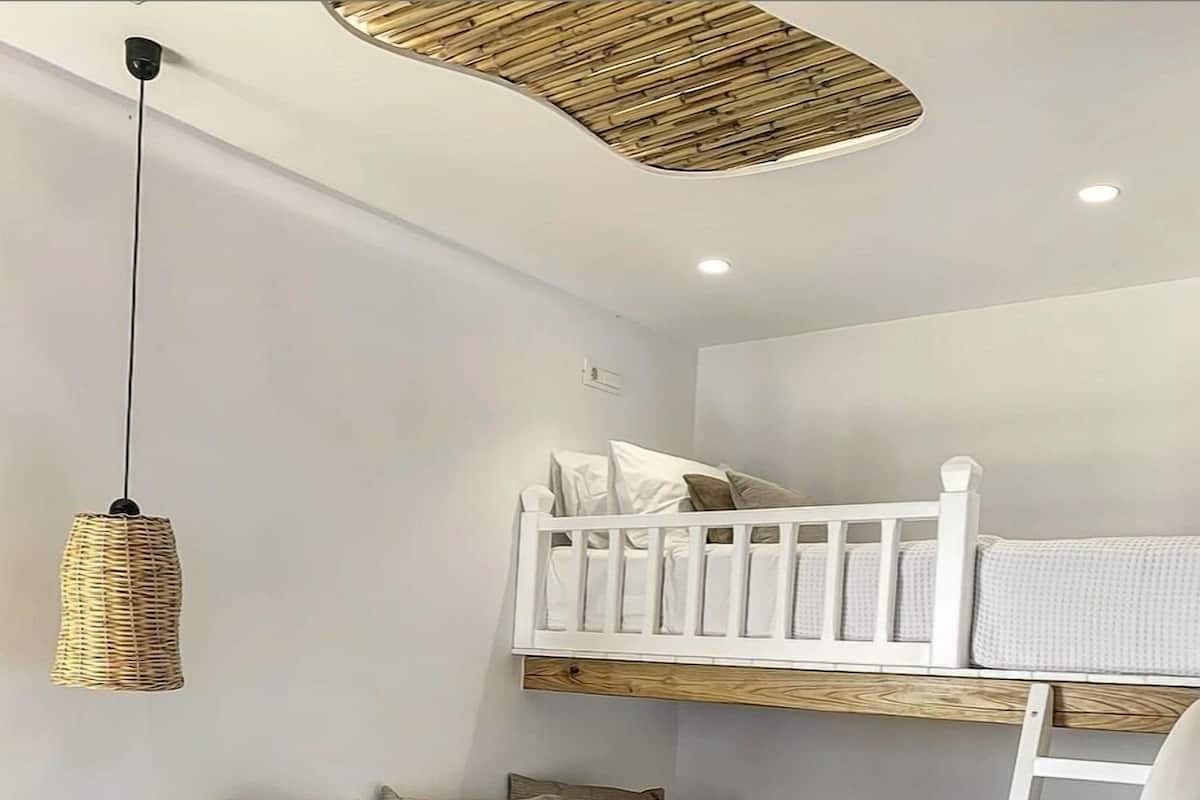 A bunk bed with a white frame, a sofa with cushions, a woven pendant light, and a white ladder.