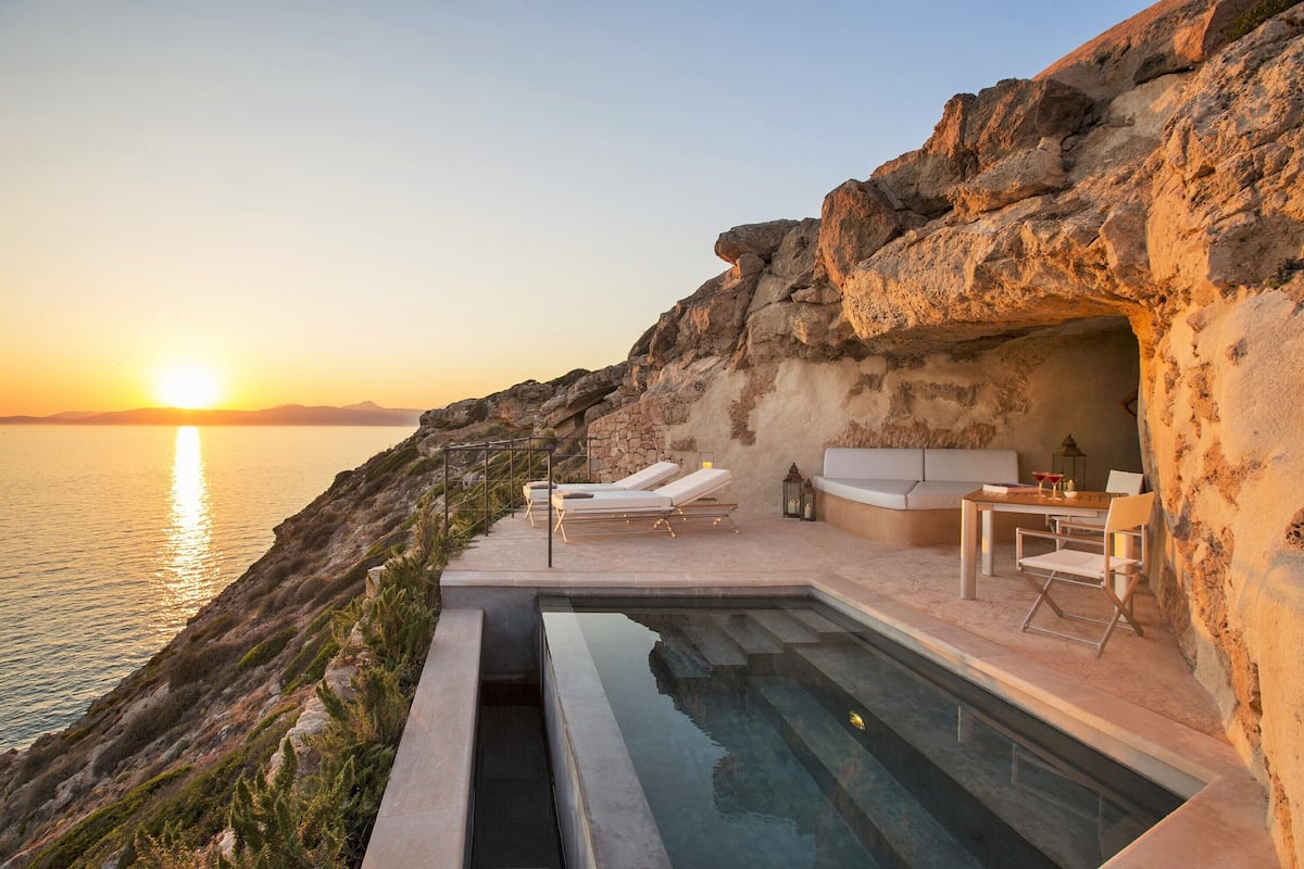 A cliffside pool with lounge chairs and a table, overlooking the sea at sunset.