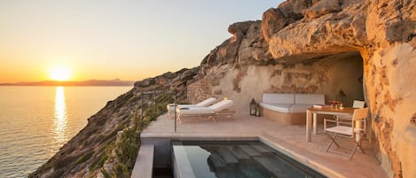 A cliffside pool with lounge chairs and a table, overlooking the sea at sunset.