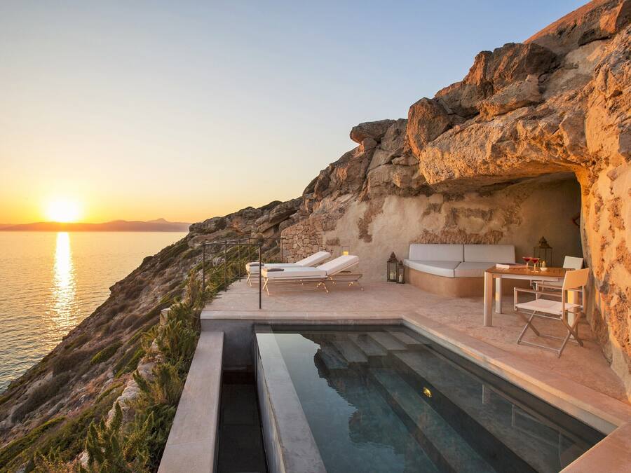 Una piscina al borde de un acantilado con sillones de descanso y una mesa, con vistas al mar al atardecer.
