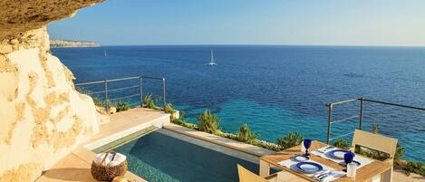An outdoor pool area with a table and chairs, a wicker basket, and a view of the sea with a sailboat in the distance.