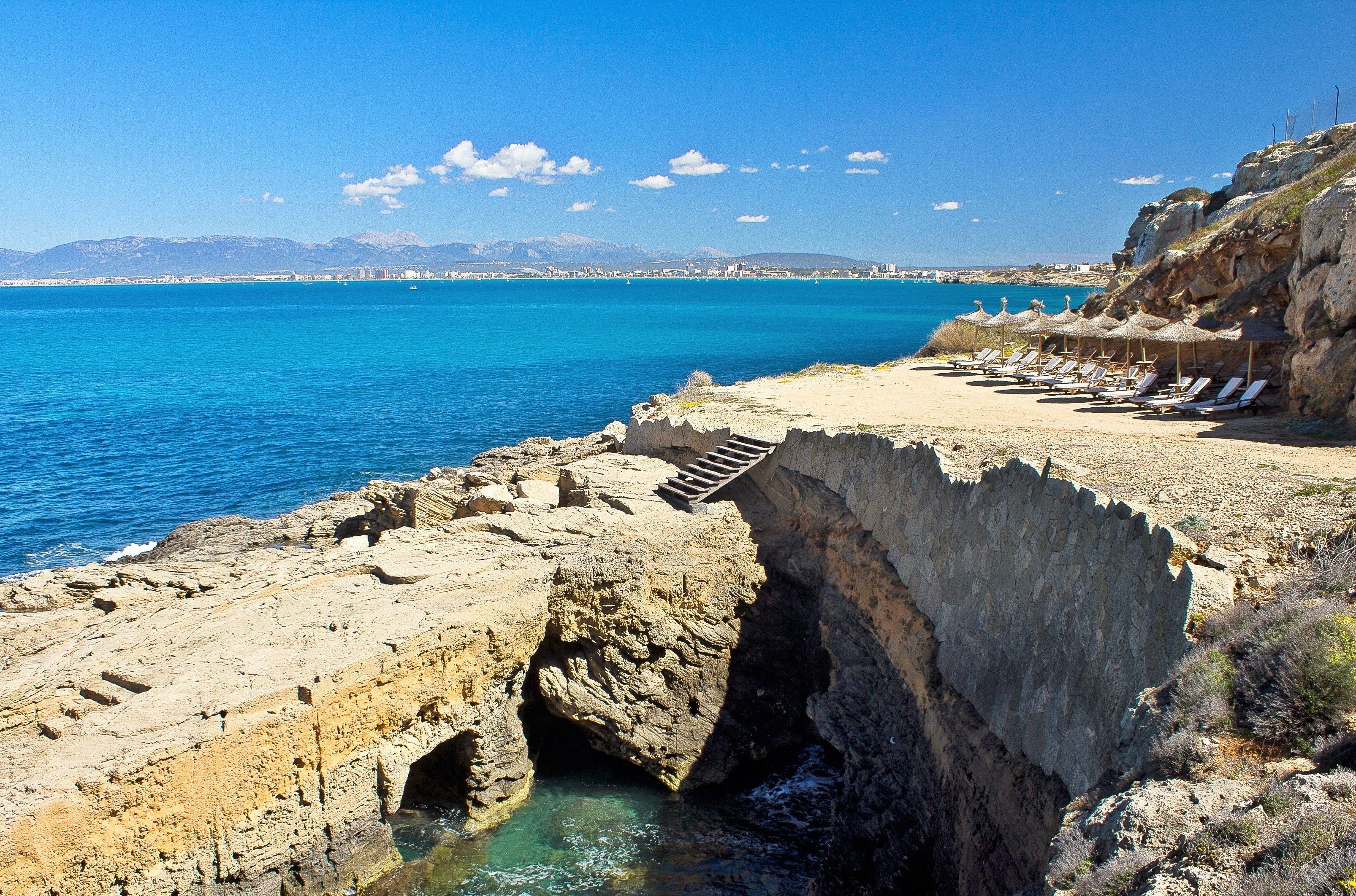 Une plage côtière bordée d’une falaise rocheuse, avec un escalier descendant jusqu’à l’eau et une rangée de transats sur le sable.